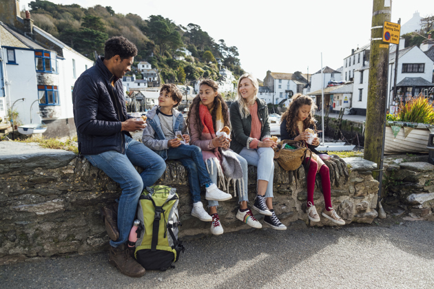Mixed family sitting on a harbour wall in a coastal village, sharing lunch and chatting together