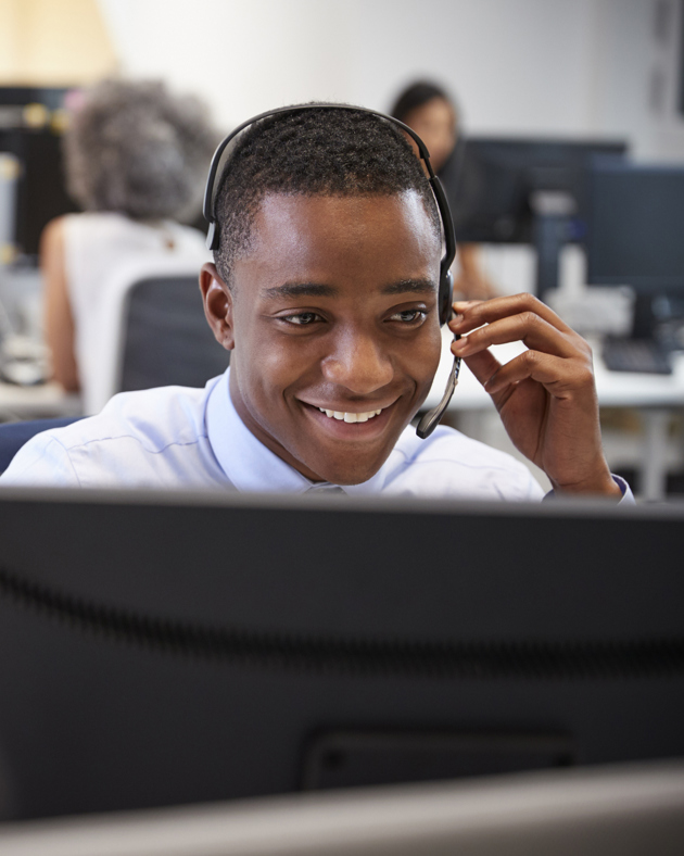 A smiling man wearing a headset is working at a computer in a modern, open-plan office.