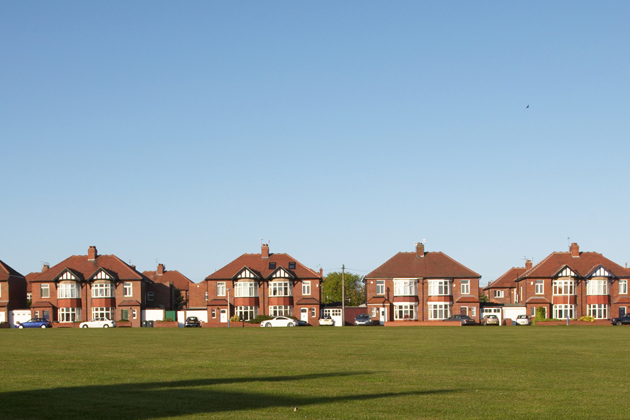 A wide row of suburban brick houses with red-tiled roofs lines the background of the image, facing a large open grassy field in the foreground.