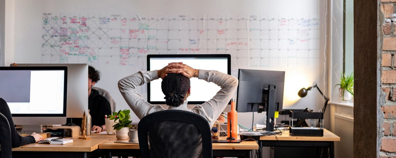 View from behind of a woman sitting at an office desk with her hands on her head
